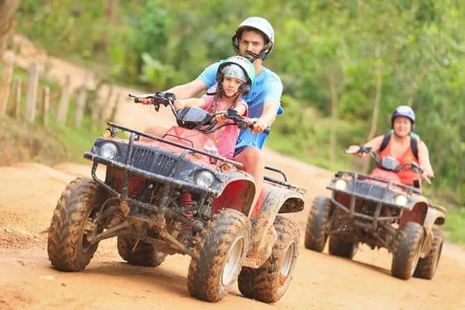 بالغ وفتاة على دراجة ATV واحدة في مسار موحل، وخلفهما راكب آخر