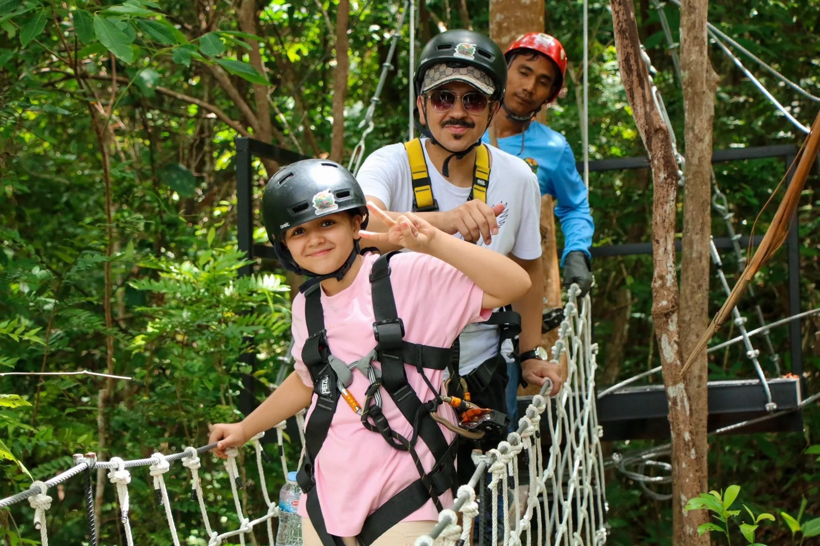 A girl and an adult in helmets and harnesses on a rope bridge, accompanied by a staff member